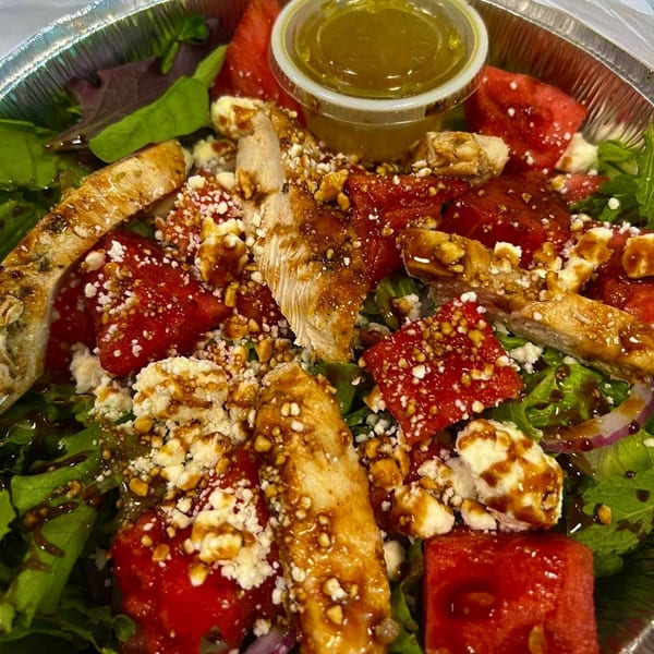 A deli salad with grilled chicken, watermelon, feta cheese, and dressing in a foil container.