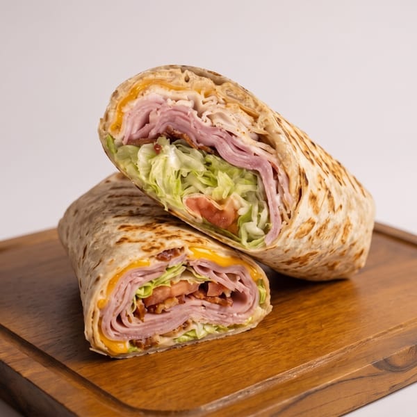 A grilled wrap, cut in half, revealing ham, cheese, lettuce, tomato, and bacon on a wooden board.
