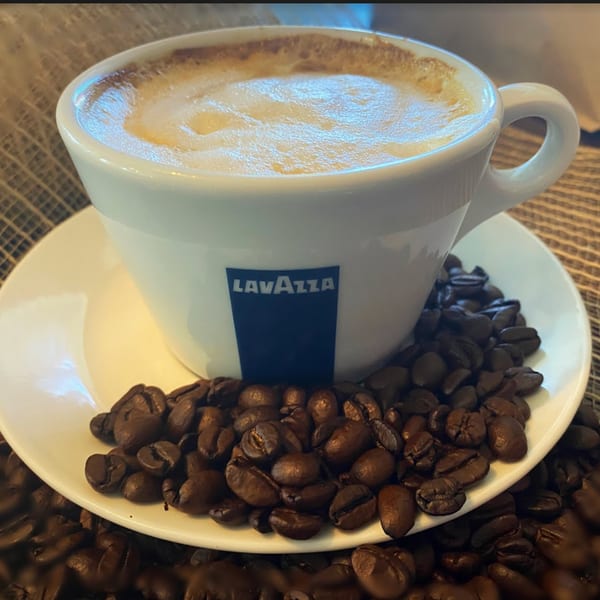 A Lavazza cappuccino in a white cup and saucer, surrounded by dark roasted coffee beans.