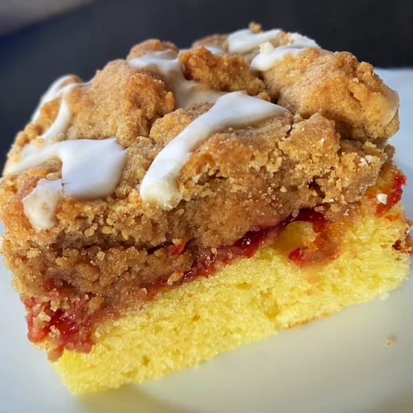 A close-up of a coffee cake slice with crumb topping, white icing, and a fruit filling.